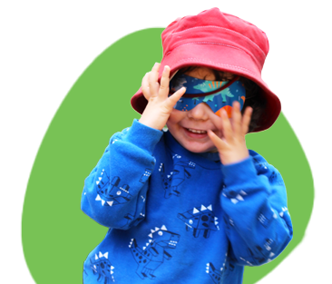 A young child wearing a red hat and blue dinosaur-themed sweatshirt, smiling and playing with a colourful mask, standing in front of a green backdrop.