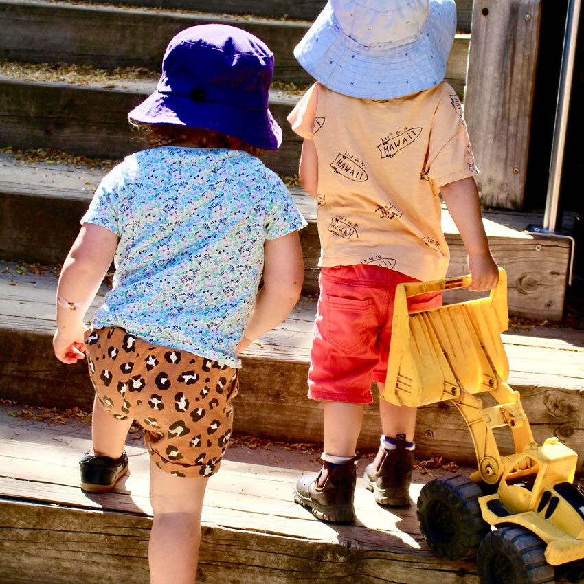 Two toddlers wearing hats and colourful outfits, climbing wooden stairs while carrying a yellow toy truck.